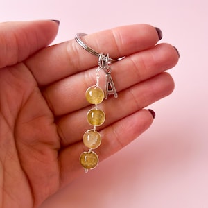 Puede incluir: Un llavero plateado con un dije de la letra 'A' y una cadena de cuatro cuentas de citrino amarillo.