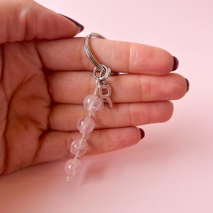 Puede incluir: Un llavero de plata con un dije de cristal rosa y un dije de letra 'R' de plata. El llavero está sostenido en una mano.