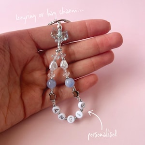 May include: A light blue and white beaded keyring with a silver charm and the name "ANGE" spelled out in white beads. The keyring is held in a hand and the text "personalised" is written on the image.