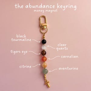 May include: A gold-toned metal keyring featuring a series of six spherical beads labeled as black tourmaline, clear quartz, tigers eye, carnelian, citrine, and aventurine. The text reads the abundance keyring money magnet with arrows pointing to each bead.