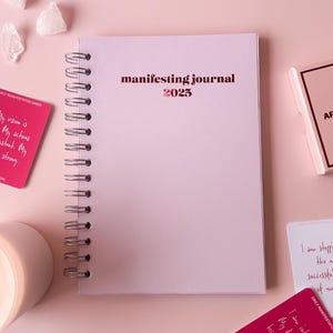 Può includere: Un diario rilegato a spirale rosa con il testo "manifesting journal 2025" sulla copertina. Il diario è circondato da altri oggetti, tra cui una carta rosa e bianca con il testo "Daily Affirmation Cards" e una carta rosa e bianca con il testo "I am stepping into the most successful decade of my life."