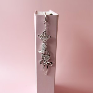 Puede incluir: Charm plateado con un diseño de planeta, bota de vaquero, corazón y lazo. El charm está suspendido de un pequeño aro y cuelga frente a un libro rosa. El charm está detallado con pequeños adornos brillantes.