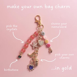 May include: A gold bag charm with various customizable elements. The charm includes pink crystal beads, letter beads, and several gold charms, including a heart-shaped lock, a starfish, and a blue heart-shaped gem. Text on the image says "make your own bag charm".