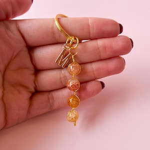 Puede incluir: Llavero dorado con un dije con la letra 'M' y una cadena de cuatro cuentas de ágata naranja y blanca.