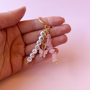 May include: A gold keychain with a white beaded charm that spells "LEGEND" and a pink rose quartz charm with two gold butterfly charms.