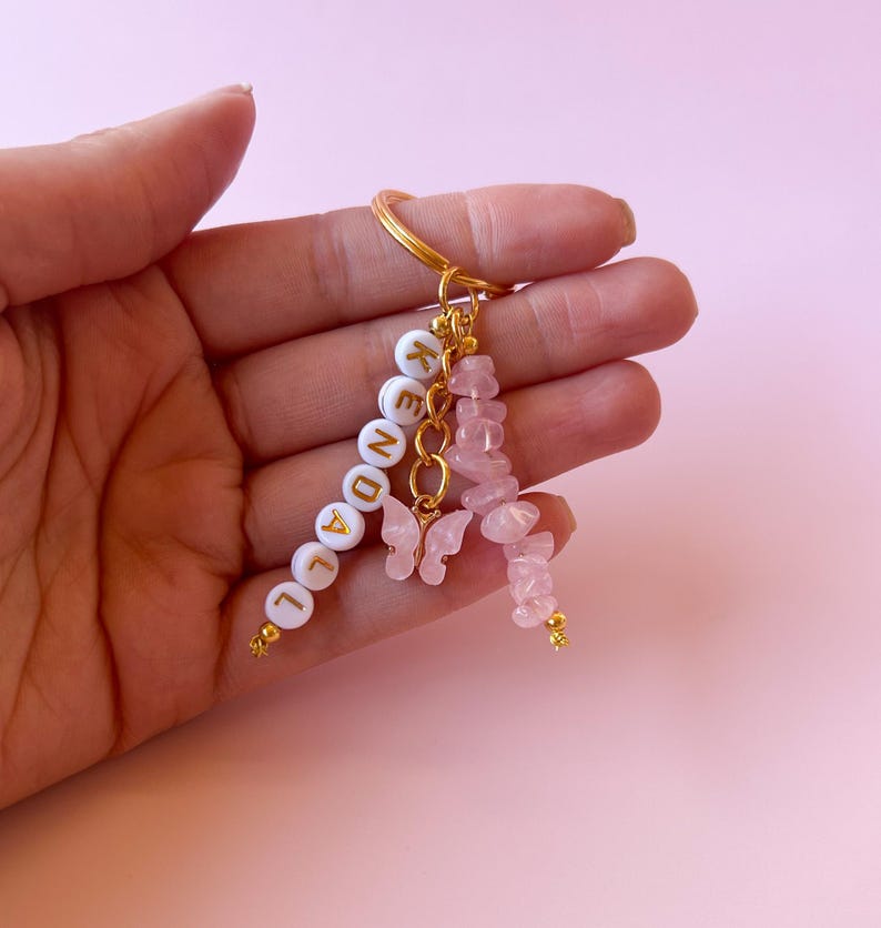 May include: A gold keychain with a white beaded charm that spells "LEGEND" and a pink rose quartz charm with two gold butterfly charms.