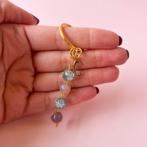 Puede incluir: Llavero dorado con un dije con la letra 'C' y una cadena colgante de cuatro cuentas redondas translúcidas de colores.