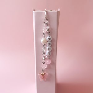 May include: A pink and white beaded bookmark with the name "RHEA" spelled out in white beads. The bookmark features a pink butterfly charm and a pearl bead.