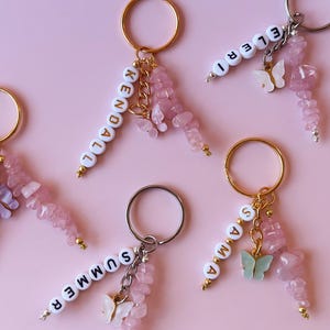 May include: Five pink and white keychains with gold rings. Each keychain has a name spelled out in white letters and a pink butterfly charm. The names are Kendall, Eleri, Summer, Anna, and a fifth name that is not visible in the image.