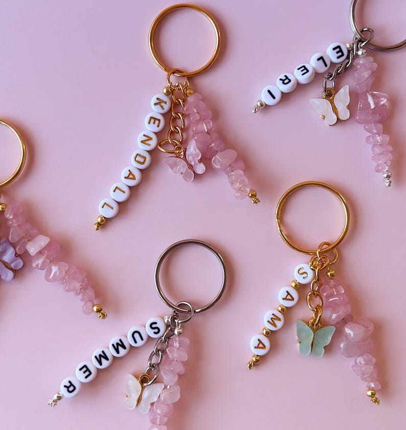 May include: Five pink and white keychains with gold rings. Each keychain has a name spelled out in white letters and a pink butterfly charm. The names are Kendall, Eleri, Summer, Anna, and a fifth name that is not visible in the image.