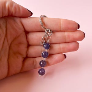 Puede incluir: Un llavero plateado con un aro grande y la letra "S" adjunta. El llavero presenta cuatro cuentas moradas ensartadas en un alambre. Las cuentas son de un tono morado intenso y tienen una apariencia suave y pulida.