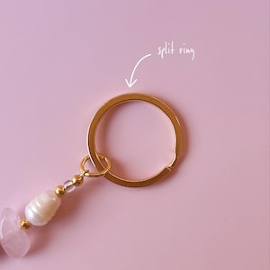 May include: A gold split ring keychain with a pink and white beaded charm. The charm features a pink crystal, a white pearl, and clear beads.