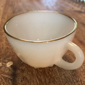 Op de afbeelding: Witte theekop van keramiek met een gouden rand. De kop heeft een ronde vorm en een handvat.