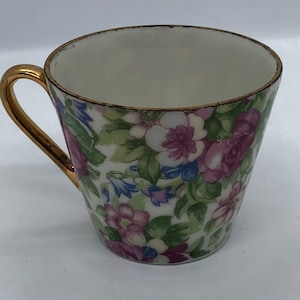 Puede incluir: Una taza de té blanca con un borde y un asa dorados. La taza está decorada con un patrón floral en tonos de rosa, morado, azul y verde.