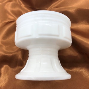 May include: A white milk glass pedestal bowl with a geometric design. The bowl has a wide, round top and a tall, slender base.