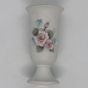 Puede incluir: Un jarrón de cerámica blanco con un diseño floral. El jarrón tiene una rosa rosa con hojas verdes y flores azules.