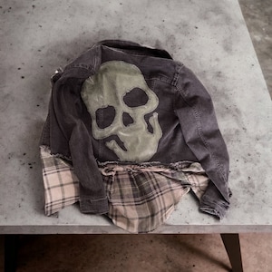 May include: A dark denim jacket with a green skull design on the back. The jacket is folded and laid on a gray concrete surface.