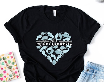 May include: Black t-shirt with a blue heart-shaped design featuring multiple manatee illustrations. The text "I'm a Manatee-aholic" is written across the heart.