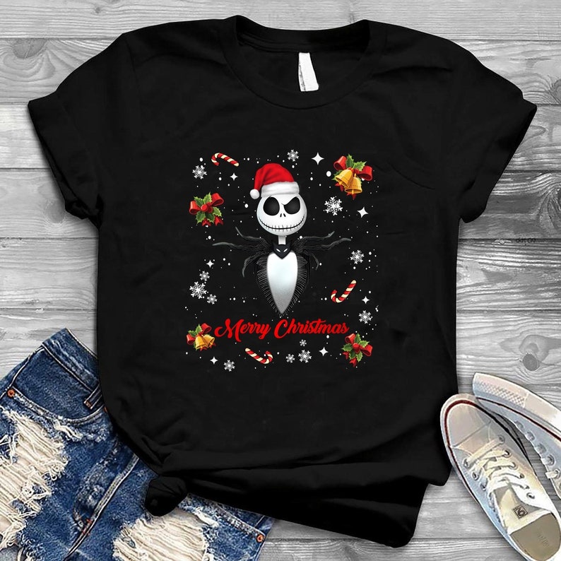 Op de afbeelding: Zwart T-shirt met een witte en zwarte illustratie van Jack Skellington uit The Nightmare Before Christmas, die een kerstmanmuts draagt. De illustratie is omringd door rode en witte pepermuntstokken, rode strikken en gouden bellen. De tekst "Merry Christmas" is in rood onder de illustratie gedrukt.
