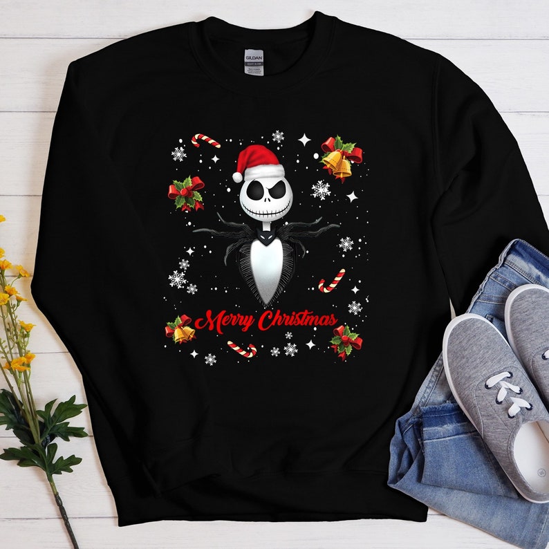 Op de afbeelding: Zwart sweatshirt met een witte en zwarte illustratie van Jack Skellington uit The Nightmare Before Christmas, die een kerstmanmuts draagt. De tekst "Merry Christmas" is onder de illustratie gedrukt. De achtergrond is zwart met witte sneeuwvlokken en snoepstokken.