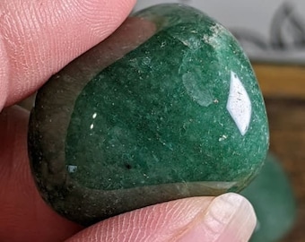 May include: A polished, tumbled green aventurine stone with a smooth, glossy surface and natural mineral inclusions. The stone displays varying shades of deep and forest green with subtle crystalline textures visible under the light.