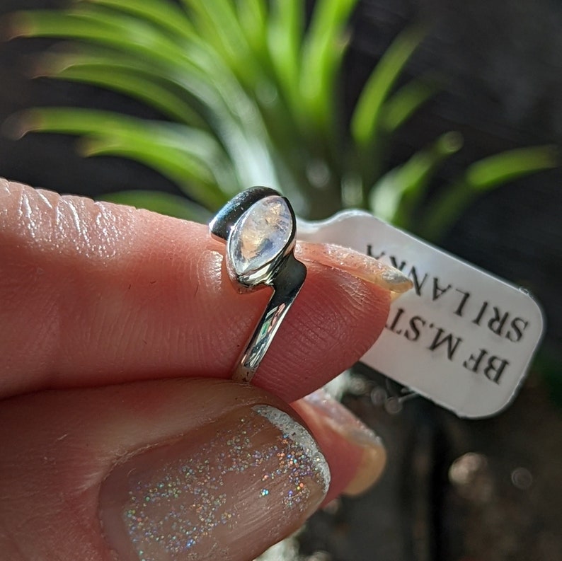 May include: A silver ring with a moonstone set in an oval shape. The ring is on a finger and the text on the tag reads "BF M'ST SRI LANKA".