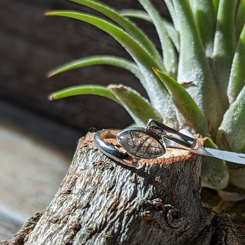 Op de afbeelding: Een zilveren ring met een donkergrijze ovale steen gezet in een zilveren band. De ring ligt op een stuk hout.