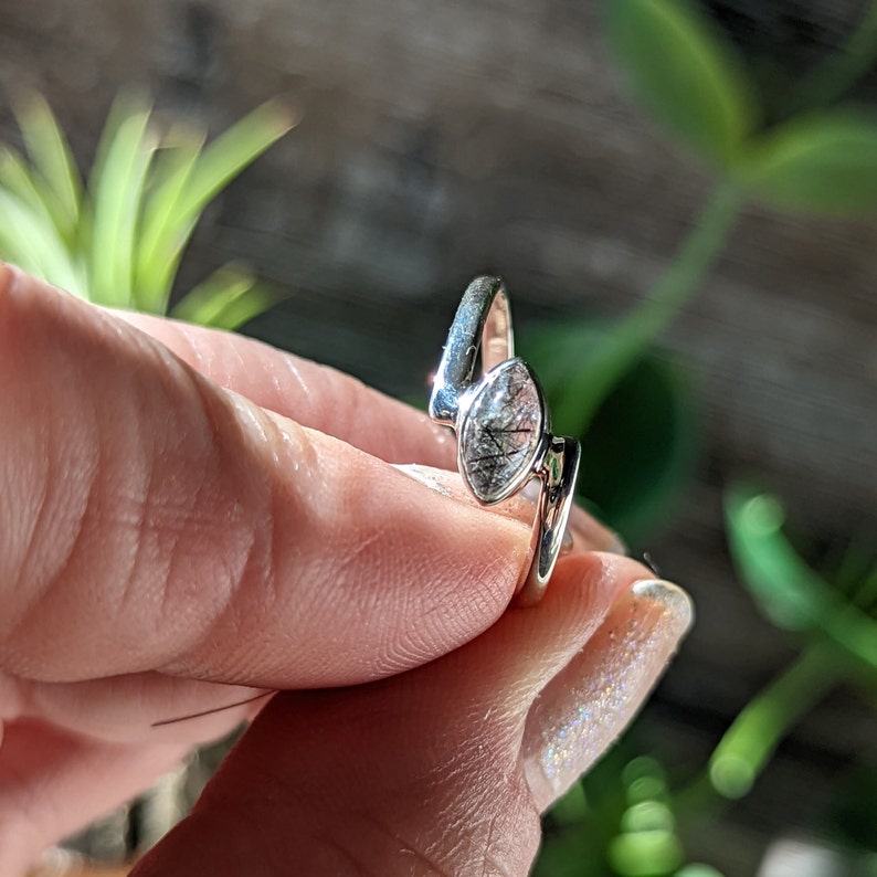 Op de afbeelding: Een zilveren ring met een heldere, ovale edelsteen met zwarte insluitsels. De ring is gezet op een zilveren band met een gebogen ontwerp.