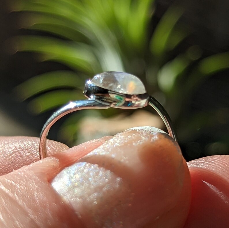 May include: A silver ring with a clear, iridescent gemstone. The ring is set on a finger.