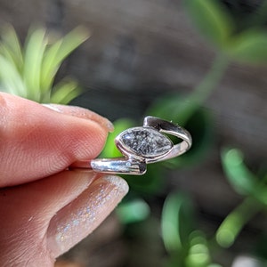 Op de afbeelding: Een zilveren ring met een markiesvormige, heldere steen met zwarte vlekken. De steen is gezet in een eenvoudige band met een gebogen ontwerp.