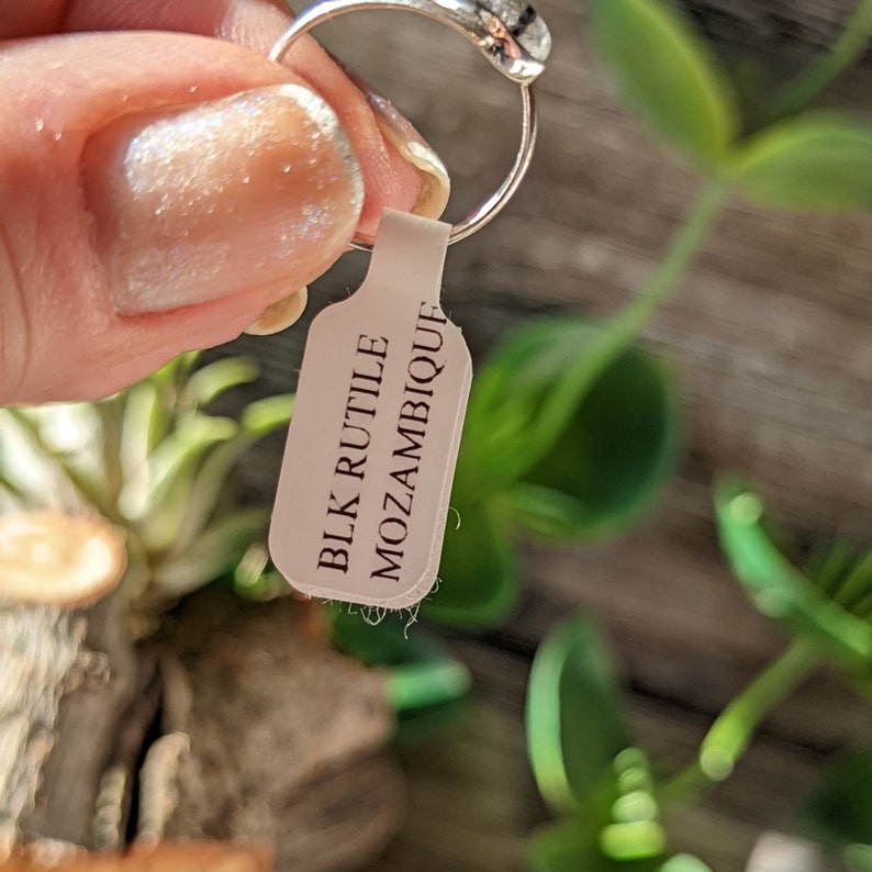 Op de afbeelding: Een zilveren ring met een zwarte rutiel steen uit Mozambique. De ring ligt op een wit label met de tekst "BLK RUTILE MOZAMBIQUE" erop.