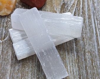 May include: Four white selenite sticks on a wooden surface. The sticks are smooth and have a natural, striated texture.