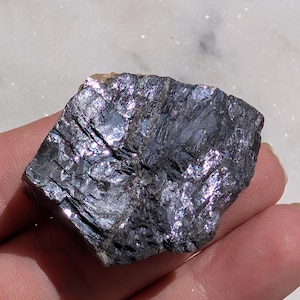 May include: A dark, iridescent mineral specimen with a layered, crystalline structure. The stone has a deep, charcoal color with shimmering, reflective surfaces. The mineral is held in a hand, showcasing its natural form and texture.