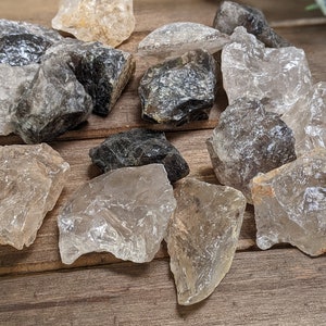 May include: A collection of raw, clear quartz crystals with smoky quartz inclusions. The crystals are various sizes and shapes, with some having a rough, natural texture.