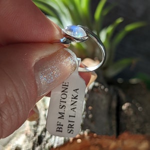 May include: A silver ring featuring a white moonstone gemstone. The ring is displayed on a white tag that reads "BF M.STONE SRI LANKA".