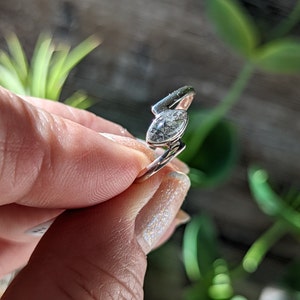 Op de afbeelding: Een zilveren ring met een heldere, marquise-vormige edelsteen met zwarte insluitsels. De ring is gezet op een zilveren band.
