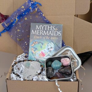 May include: A brown cardboard box filled with a variety of items, including a deck of oracle cards titled "Myths & Mermaids Oracle of the Water", a clear crystal, a brown and gold beaded bracelet, and a small metal tin filled with colourful gemstones.