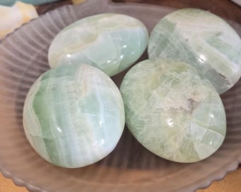 May include: Four polished, oval-shaped green stones rest on a decorative plate. The stones have a translucent quality with white and light green marbling. The plate sits on a wooden surface, with other crystals and decorative items in the background.
