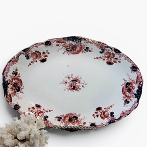 May include: An oval ceramic platter with a scalloped edge, featuring a white base decorated with reddish-brown floral patterns and dark blue accents. The design includes a central floral motif surrounded by a border of repeating flower clusters.
