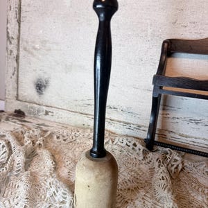 May include: A vintage wooden muddler with a black handle and a light-colored wooden base. The muddler has a rounded top and a cylindrical base. The background includes a lace cloth and a small black chair.