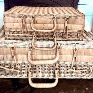 May include: Two rectangular woven wicker suitcases stacked on top of each other. Each suitcase features a light tan natural finish, a hinged lid, and a sturdy arched handle attached to the front. The weave pattern is consistent across both cases.