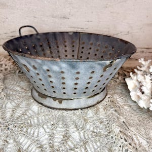Op de afbeelding: Een vintage vergiet van gegalvaniseerd metaal met een verweerde blauwgrijze afwerking. Het vergiet heeft een ronde komvorm met talrijke kleine afvoergaten en twee kleine handvatten. Het staat op een cirkelvormige basis.