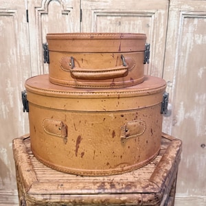 May include: Two stacked oval-shaped tan leather hat boxes with a speckled finish. The top box features a leather handle and metal side latches. The larger bottom box has matching metal latches on each side and reinforced leather handle attachments.