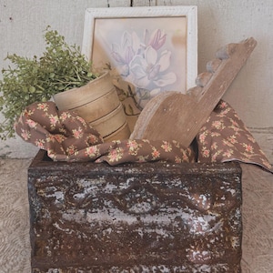 May include: A rustic, weathered metal box filled with decorative items. Inside are a framed floral print, a stack of small wooden containers, a wooden decorative piece, and fabric with a floral pattern. Greenery adds a natural touch.