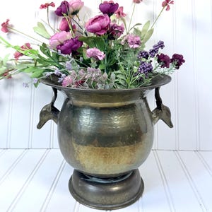 May include: A vintage brass vase with a hammered texture, featuring two decorative handles. The vase is filled with an arrangement of artificial flowers in shades of purple, pink, and green. The vase sits on a white surface.