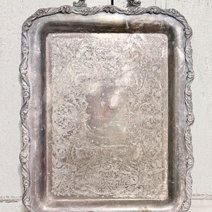 May include: A rectangular silver-colored serving tray with ornate handles and a detailed floral pattern. The tray has a decorative border and is likely made of metal. The design suggests a vintage or antique style, suitable for serving food or displaying items.