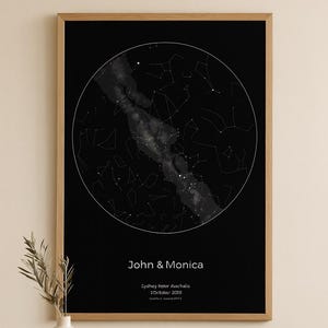May include: A framed print of a star map in black and white. The print shows a circular representation of constellations and the Milky Way. Text at the bottom reads "John & Monica" with location and date details. The frame is light brown.