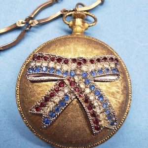Peut inclure: Un pendentif de montre de poche doré avec un nœud décoratif. Le nœud est orné de strass rouges, blancs et bleus. La montre a une surface texturée et est attachée à une chaîne dorée.