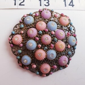 May include: A vintage brooch with a floral design. The brooch features a filigree metal frame with pastel pink, blue, and purple flower-shaped accents, each with a central rhinestone. Small, colorful beads surround the flowers.