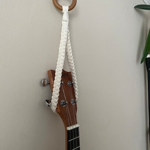 Könnte beinhalten: Ein weißer Makramee-Wandhalter mit einem Holzring, an dem eine Ukulele hängt.